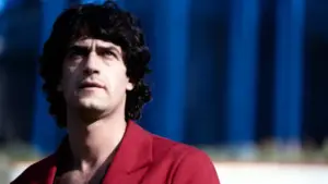 Un uomo con capelli scuri ricci guarda verso l’alto, indossando una giacca rossa.