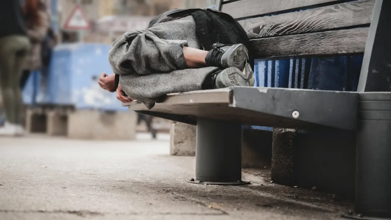 Persona accovacciata su una panchina in strada, coperta da abiti pesanti