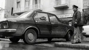 Un agente osserva un'auto danneggiata parcheggiata in strada, scena legata al caso Mattarella