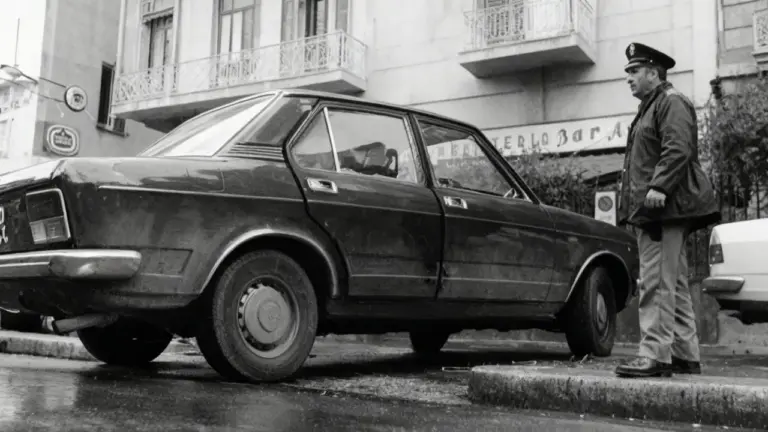 Un agente osserva un'auto danneggiata parcheggiata in strada, scena legata al caso Mattarella