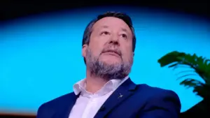 Un uomo in abito blu durante un'intervista televisiva, con sfondo blu e pianta sullo sfondo.