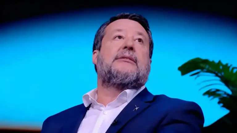 Un uomo in abito blu durante un'intervista televisiva, con sfondo blu e pianta sullo sfondo.