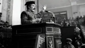 Un uomo in divisa militare parla da un podio in un'aula con altre persone armate presenti