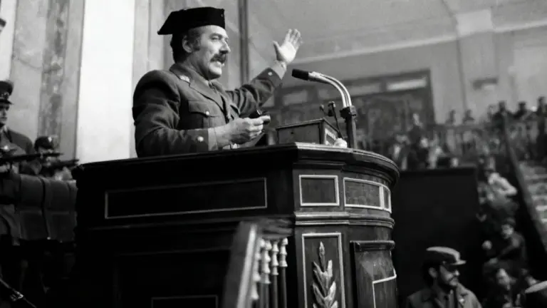 Un uomo in divisa militare parla da un podio in un'aula con altre persone armate presenti