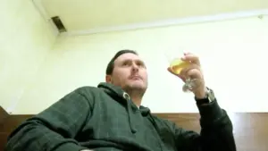 Un uomo seduto con una felpa verde scura tiene in mano un bicchiere con una bevanda chiara