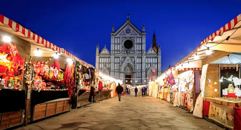 Bancarelle illuminate di un mercatino di Natale in una piazza con una chiesa storica sullo sfondo