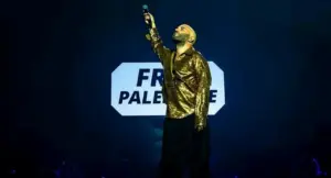 Una persona su un palco durante un concerto, con un microfono alzato e uno sfondo con scritta visibile.