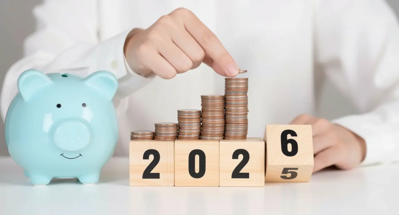 Mano che impila monete accanto a salvadanaio e blocchi con l'anno 2026