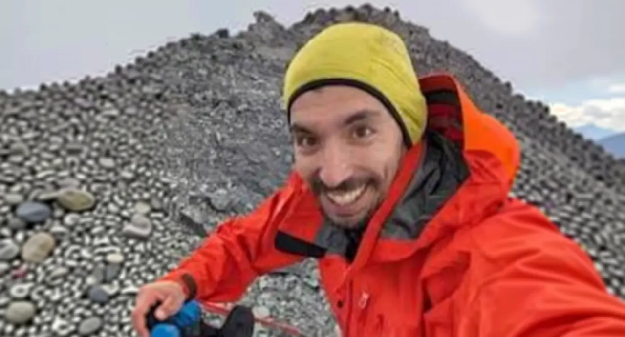 Una persona in giacca rossa e cappello giallo sorride in primo piano, con sfondo montano roccioso